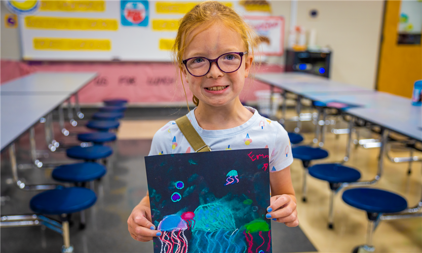 A girl with red hair and glasses holds up a piece of art